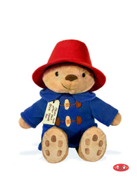A plush teddy bear dressed as Paddington Bear, wearing a red hat and blue duffel coat, seated against a plain white background with a small red logo in the bottom right corner.