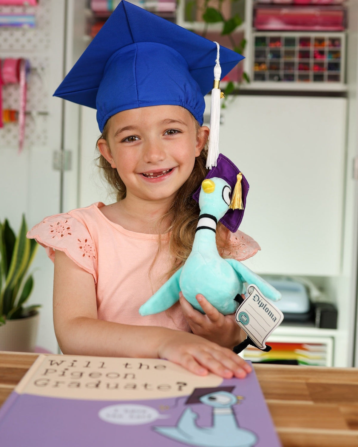 Child wearing a blue graduation cap holding a plush pigeon with a diploma, in a classroom setting.