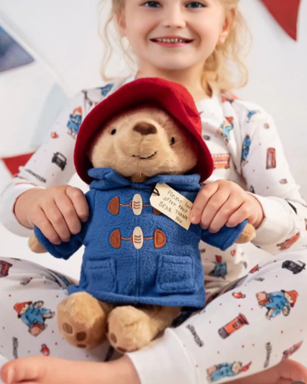 Child holding a Paddington teddy bear with a red hood and blue coat, wearing pajamas with similar design.