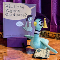 Plush pigeon toy in graduation attire with a book titled 'Will the Pigeon Graduate?' on a wooden surface.