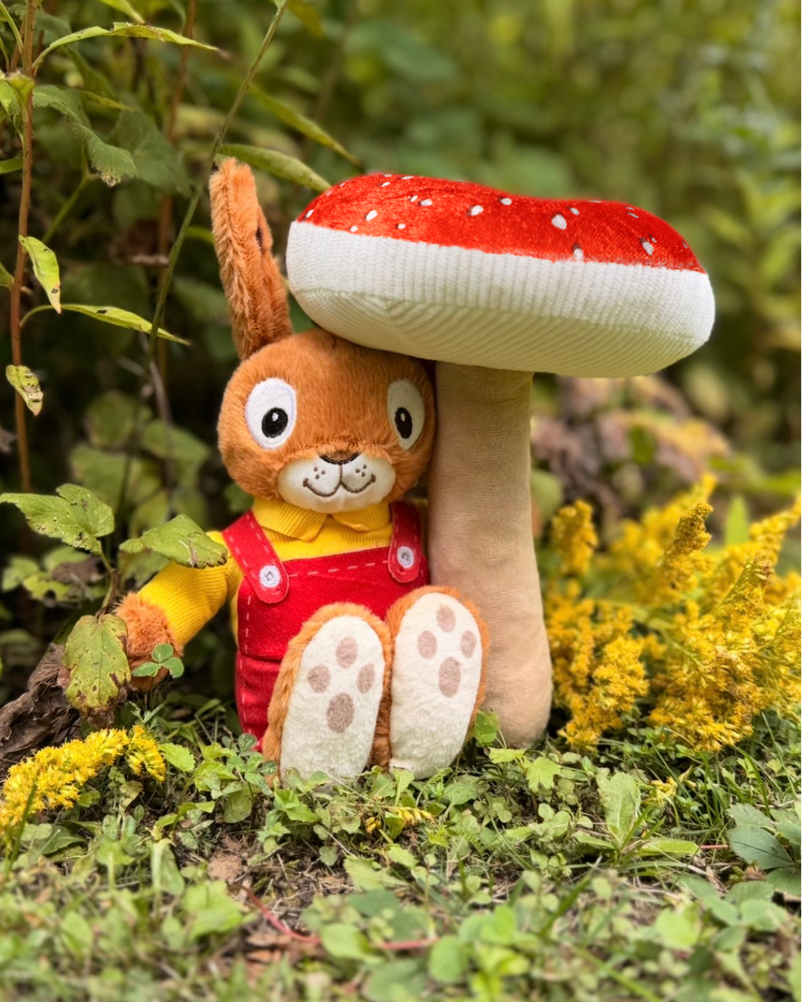 A plush toy rabbit in red overalls sitting beside a large red-and-white mushroom, placed outdoors among green leaves and yellow flowers.