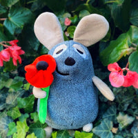 A gray plush mouse toy holding a red poppy flower, set against a background of green ivy leaves and pink flowers.